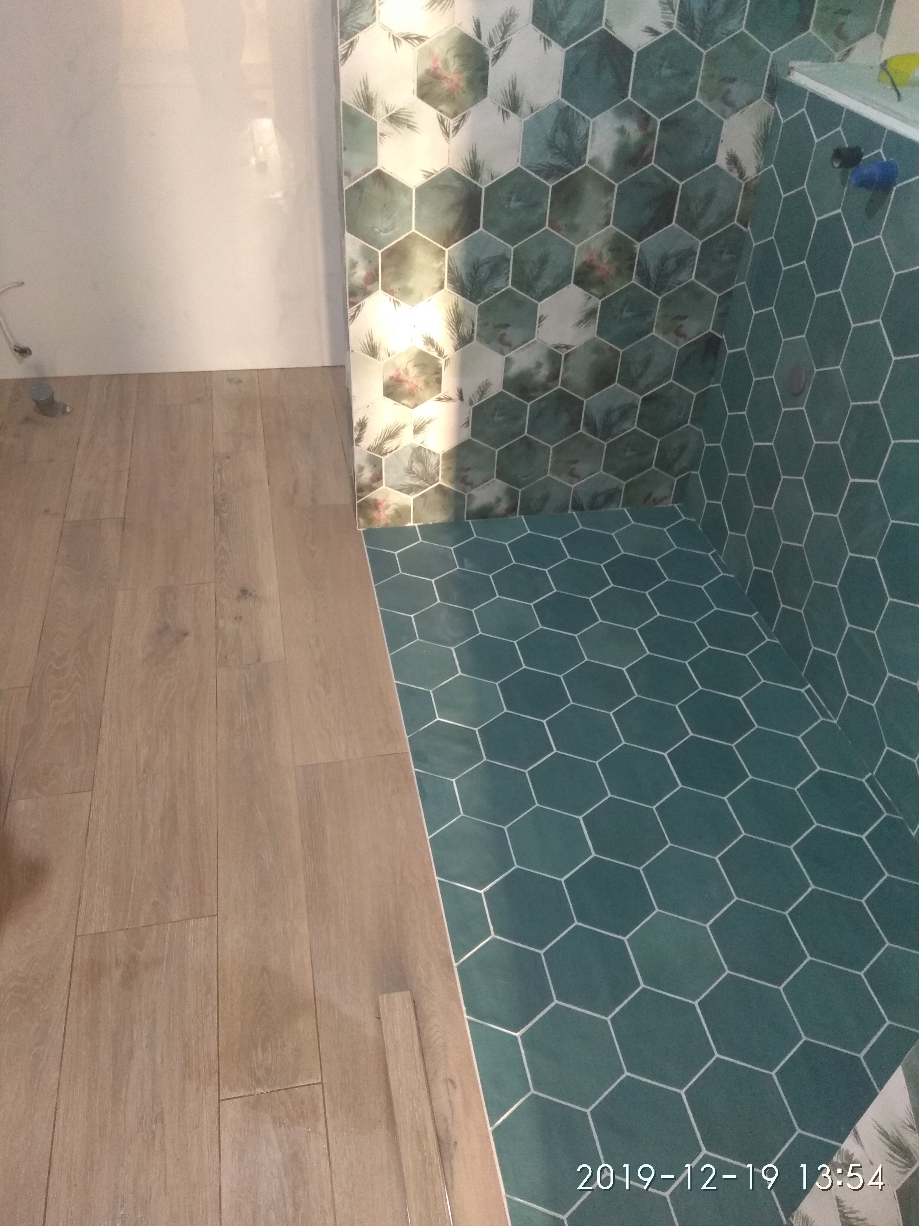 Wykończona łazienka z drewnopodobną podłogą i heksagonalnymi, turkusowymi płytkami na podłodze i ścianach, z wzorem roślinnym na części płytek ściennych.