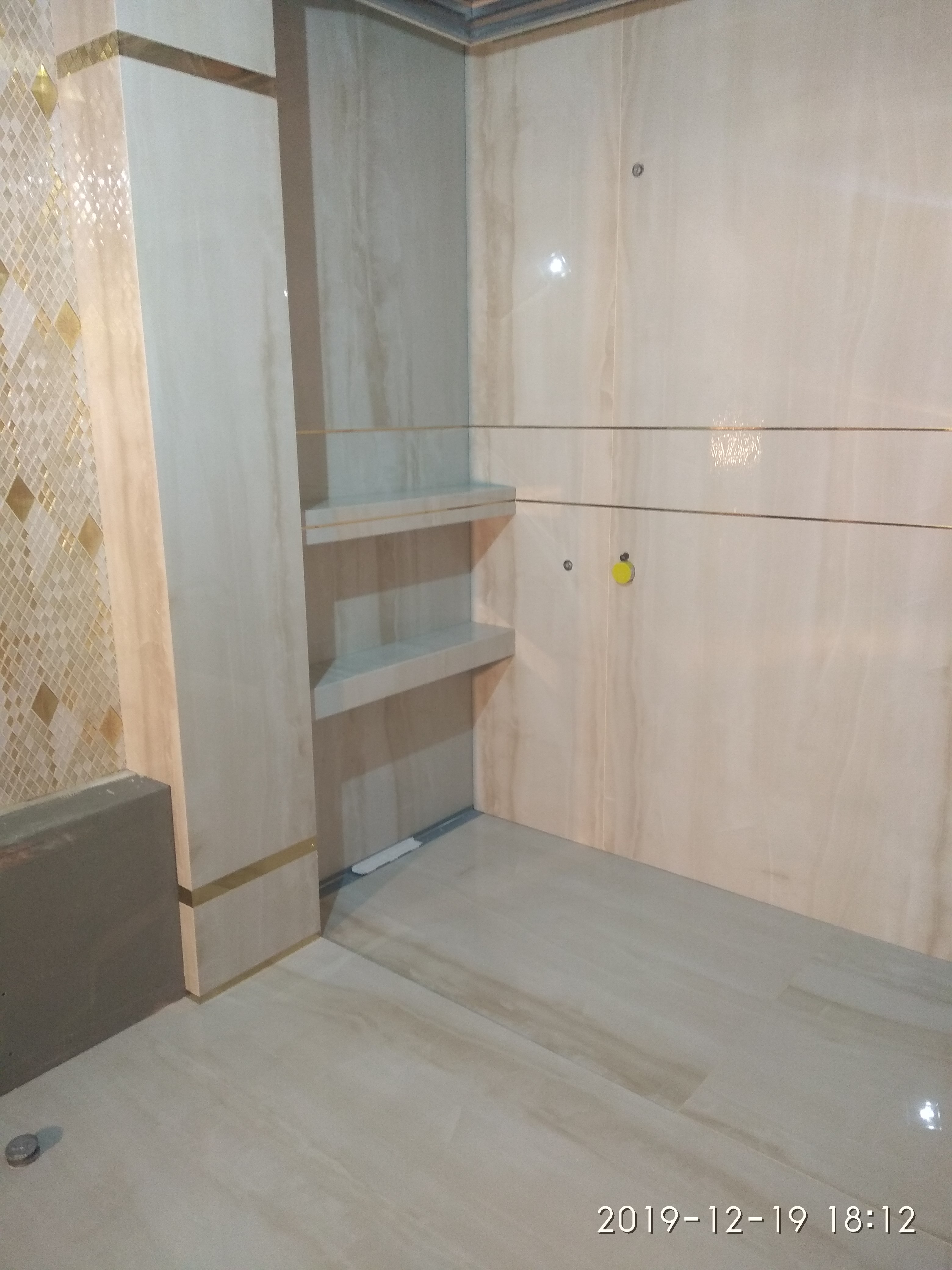 Wykończona łazienka z beżowymi płytkami imitującymi marmur, mozaika w odcieniach złota, wnęka z półkami, widoczne punkty instalacji wodnej.