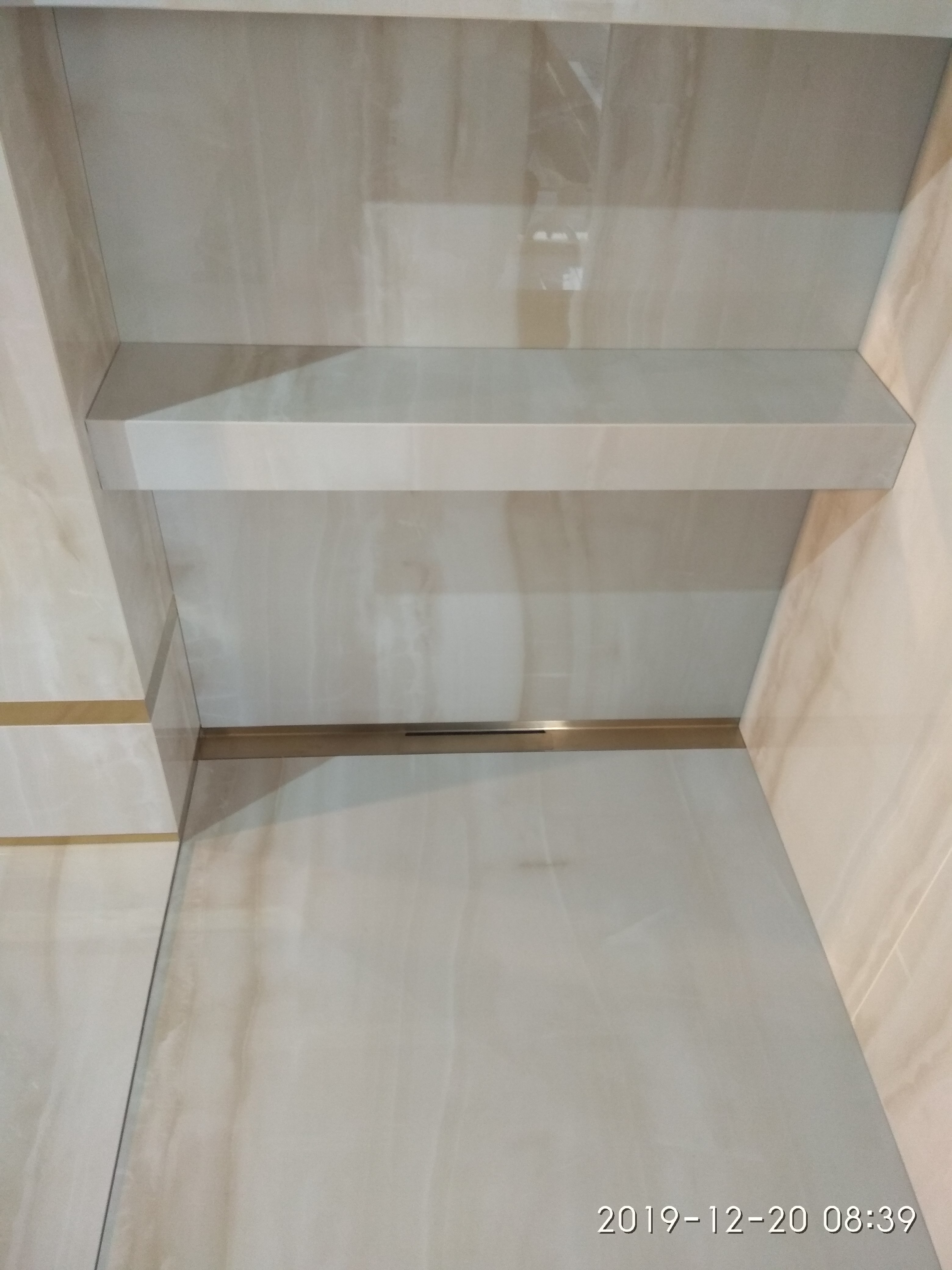 Wykończona wnęka z półką w łazience, wyłożona jasnymi płytkami imitującymi marmur, z delikatnymi złotymi akcentami.