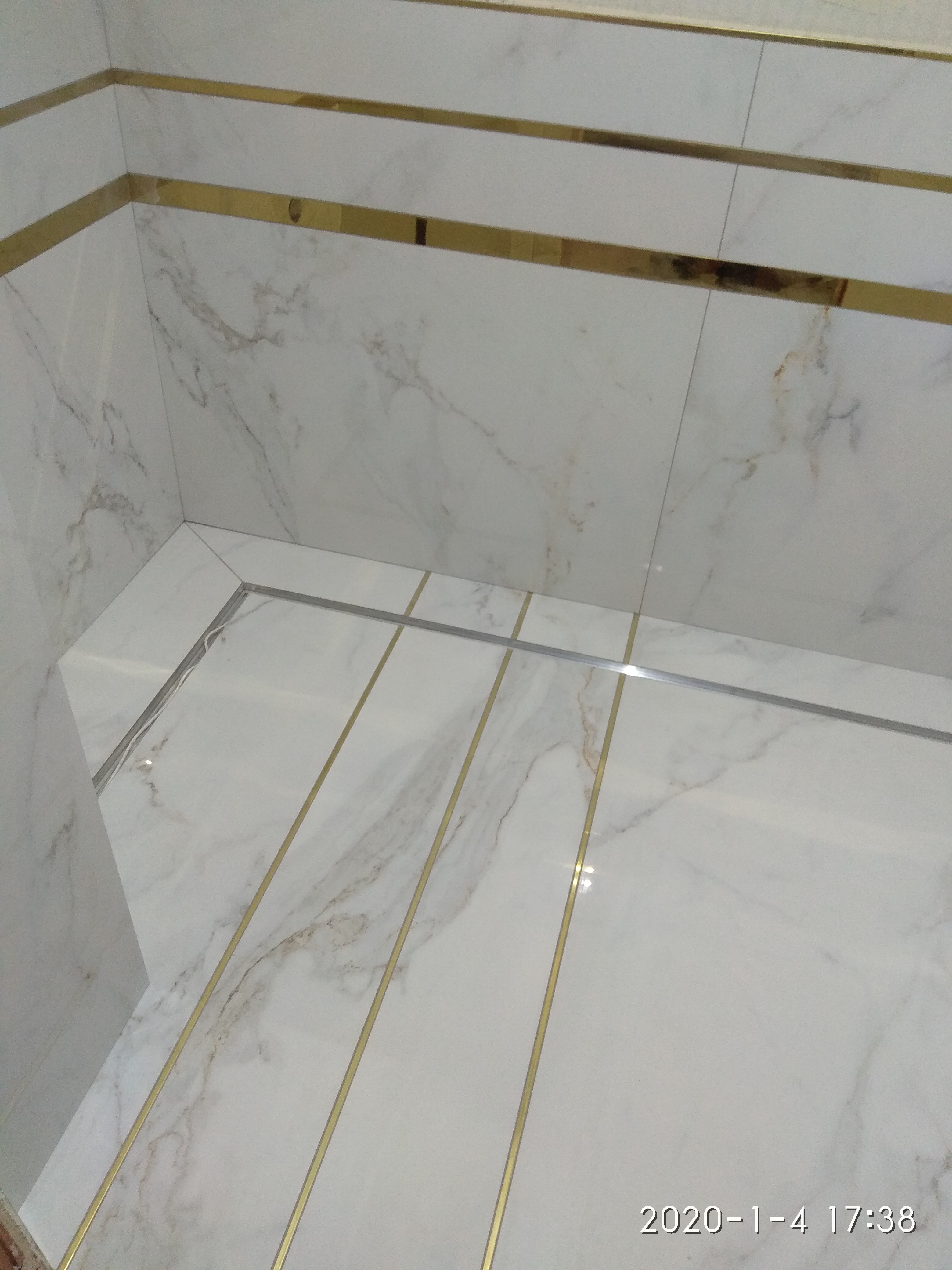 Wykończona łazienka z białymi marmurkowymi płytkami i złotymi listwami dekoracyjnymi, widok z góry na podłogę i fragment ściany.
