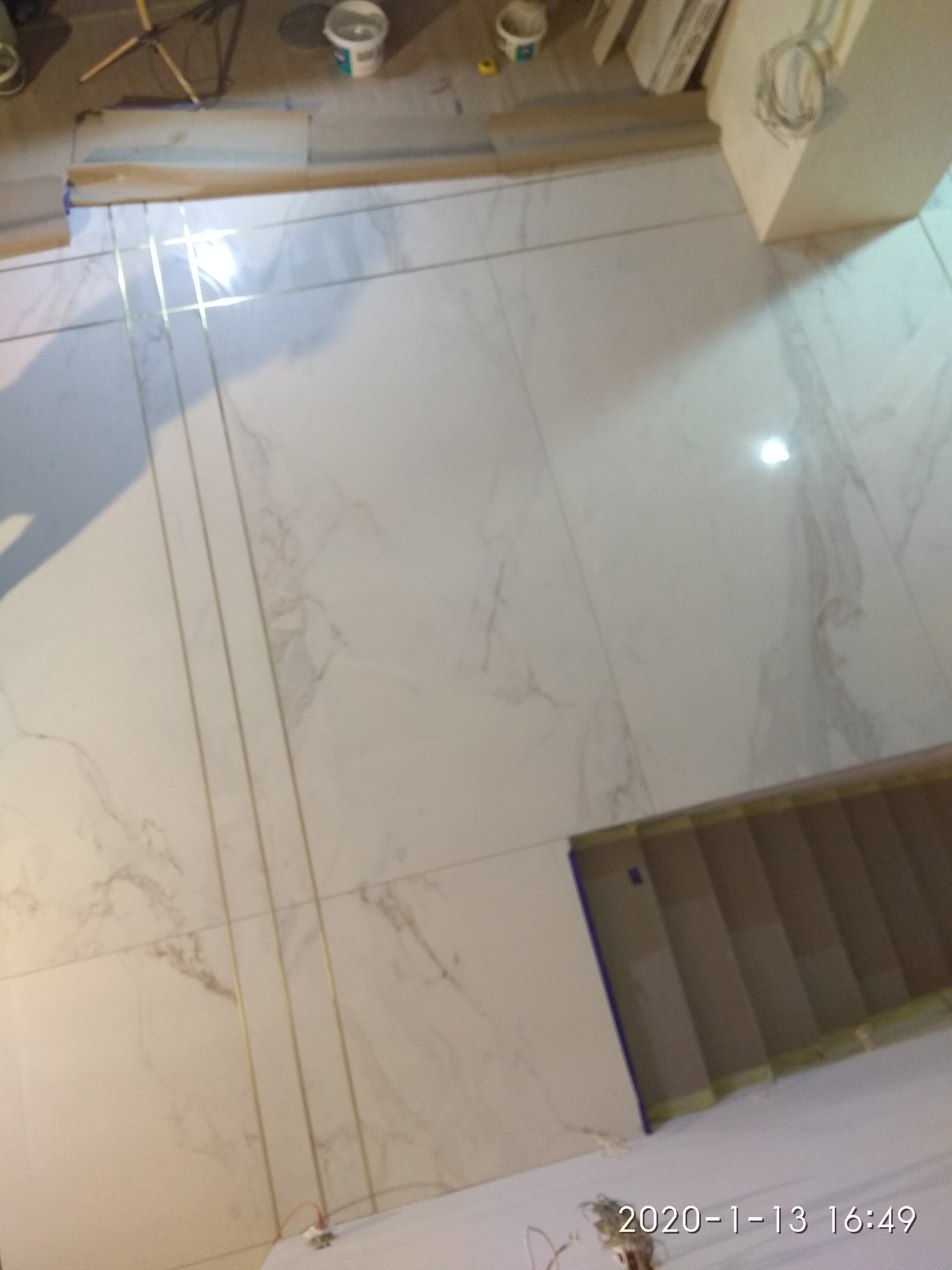 Ułożone, jasne płytki podłogowe z delikatnym marmurkowym wzorem, oddzielone złotymi listwami, z widocznymi narzędziami i materiałami budowlanymi w tle.