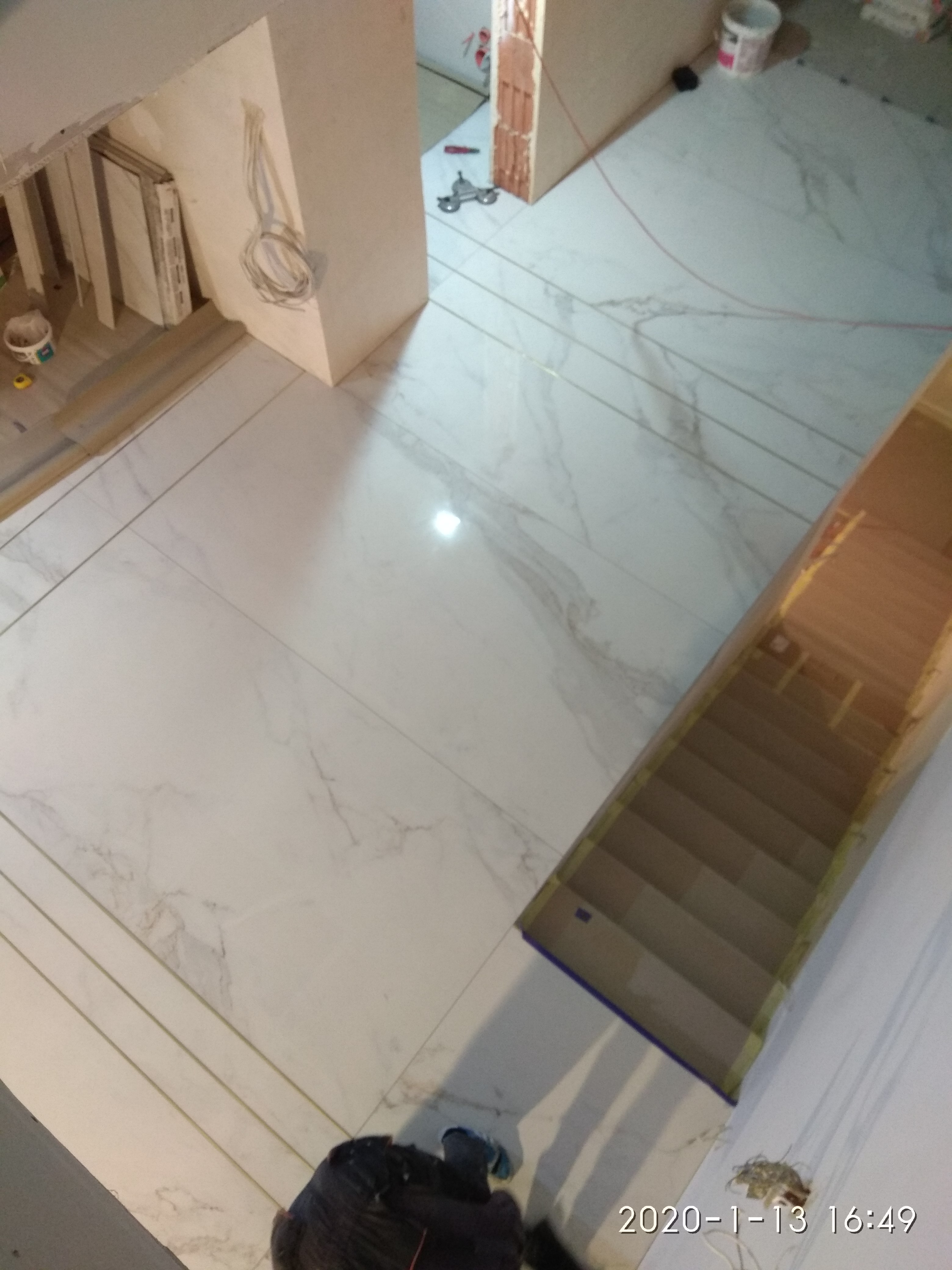 Widok z góry na białe płytki podłogowe imitujące marmur, ułożone w trakcie remontu, widoczne narzędzia glazurnicze, przycięte płytki i kable elektryczne wystające ze ściany.