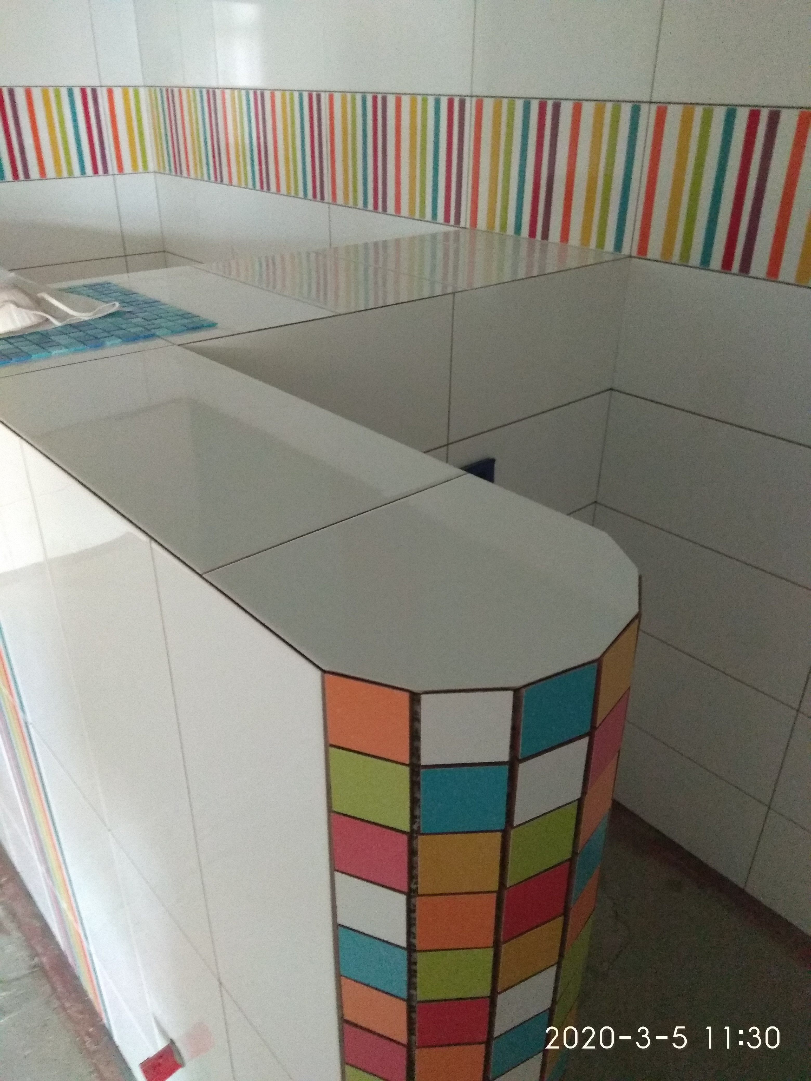 Wykończona ściana i blat z białych płytek z kolorowymi akcentami: pasem pionowych pasków na ścianie i mozaiką z kolorowych kwadratów na zaokrąglonym narożniku blatu.