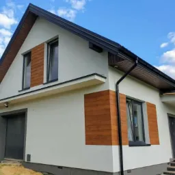 Nowoczesny dom jednorodzinny z jasną elewacją, drewnianymi akcentami na ścianach i ciemnym dachem, widok z zewnątrz w słoneczny dzień.