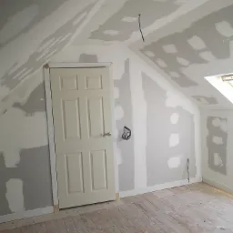 Poddasze w trakcie prac wykończeniowych: ściany i sufit z płyt kartonowo-gipsowych, szpachlowane łączenia, drewniana podłoga, widoczne przewody elektryczne, białe drzwi z ościeżnicą.