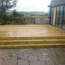 Świeżo wybudowany drewniany taras z trzema stopniami, przylegający do budynku z szarymi panelami i dużymi przesuwnymi drzwiami, na tle kamiennego muru i zieleni.