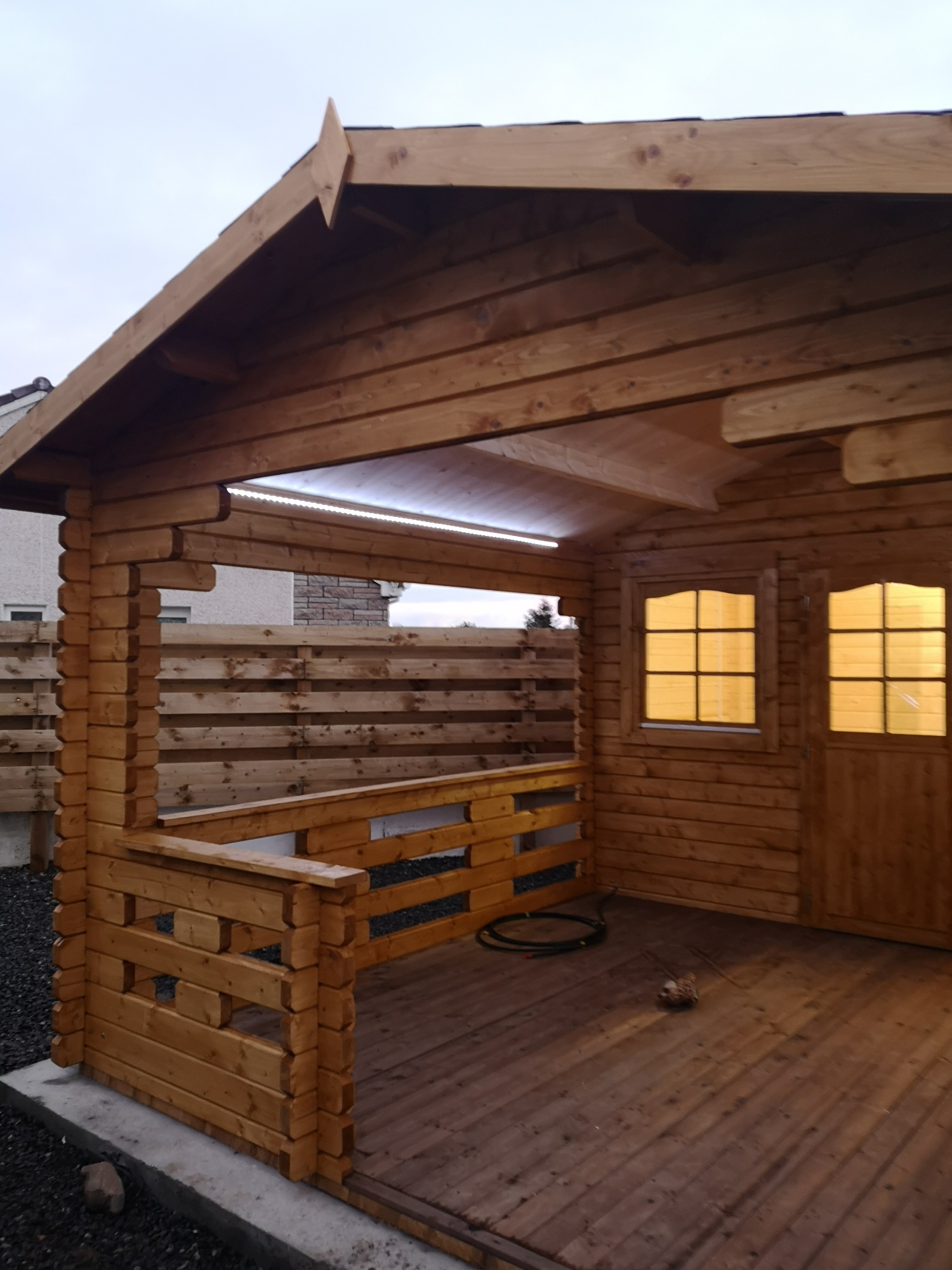 Drewniana altana z podświetleniem LED pod dachem, widok z zewnątrz na drewnianą podłogę i okno z drzwiami, czarny kabel leżący na podłodze.