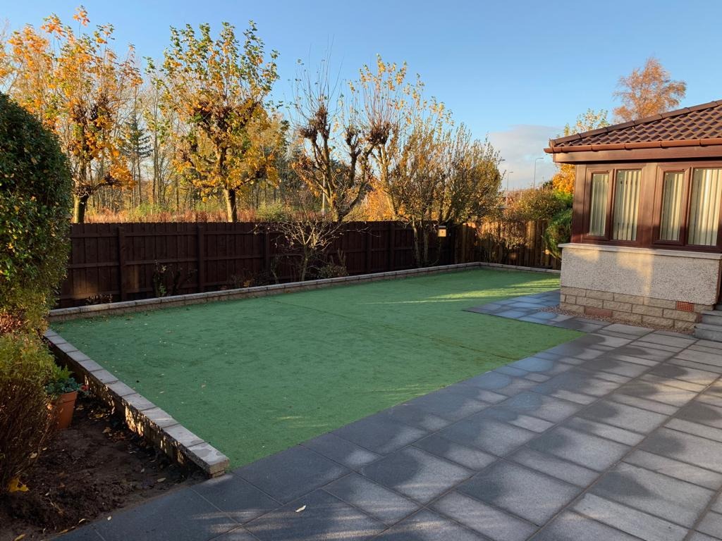 Ułożona szara kostka brukowa sąsiadująca z trawnikiem ze sztucznej trawy, otoczona niskim murkiem i brązowym drewnianym ogrodzeniem na tle jesiennego krajobrazu z drzewami.