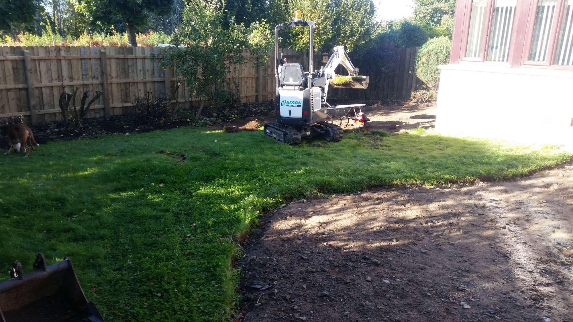 Mini koparka Nixon z łyżką pełną darni na trawniku podczas prac ziemnych w ogrodzie, widoczny fragment budynku z oknami, ogrodzenie drewniane i pies.