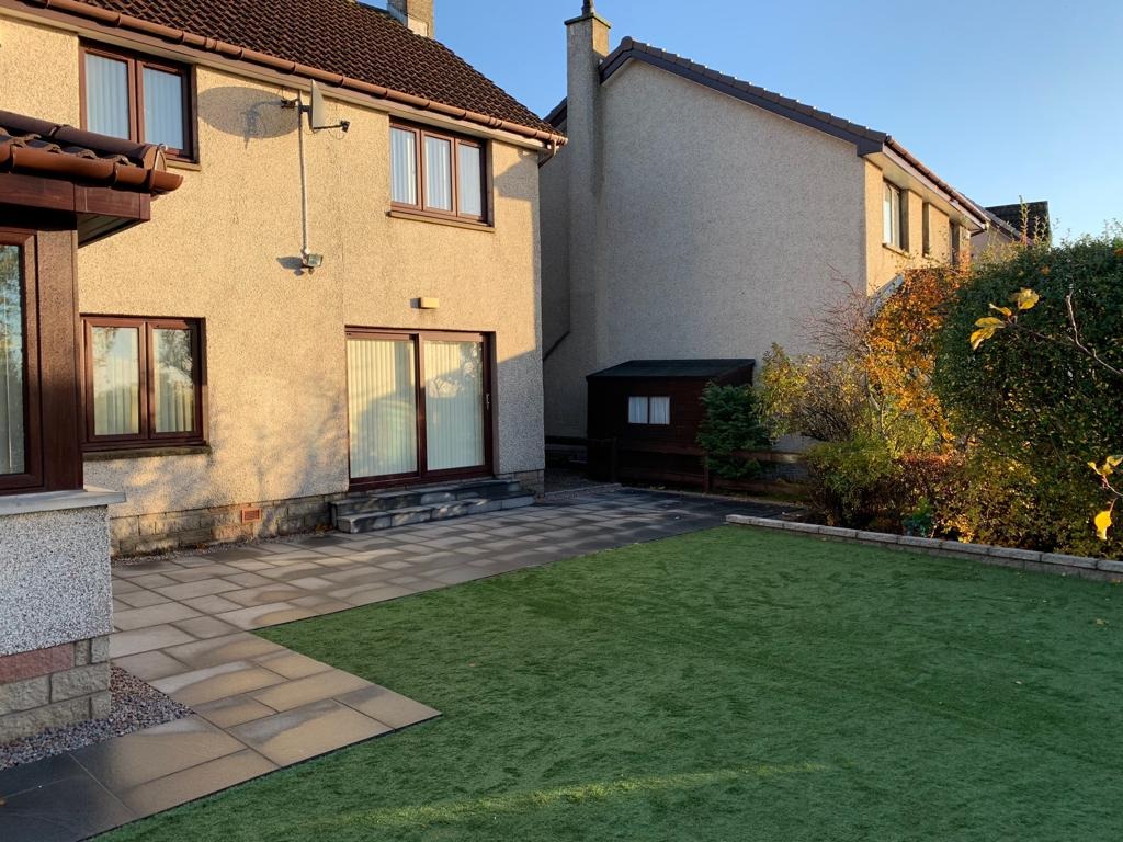 Elewacja domu z brązowymi oknami i przesuwnymi drzwiami, nowy taras z szarych płyt i trawnik z sztucznej trawy w słoneczny dzień.