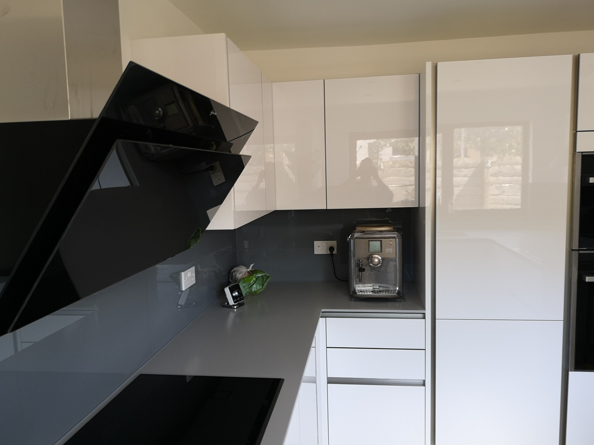 Nowoczesna kuchnia z białymi, lakierowanymi szafkami, szarym blatem i czarnym okapem, ekspres do kawy na blacie.