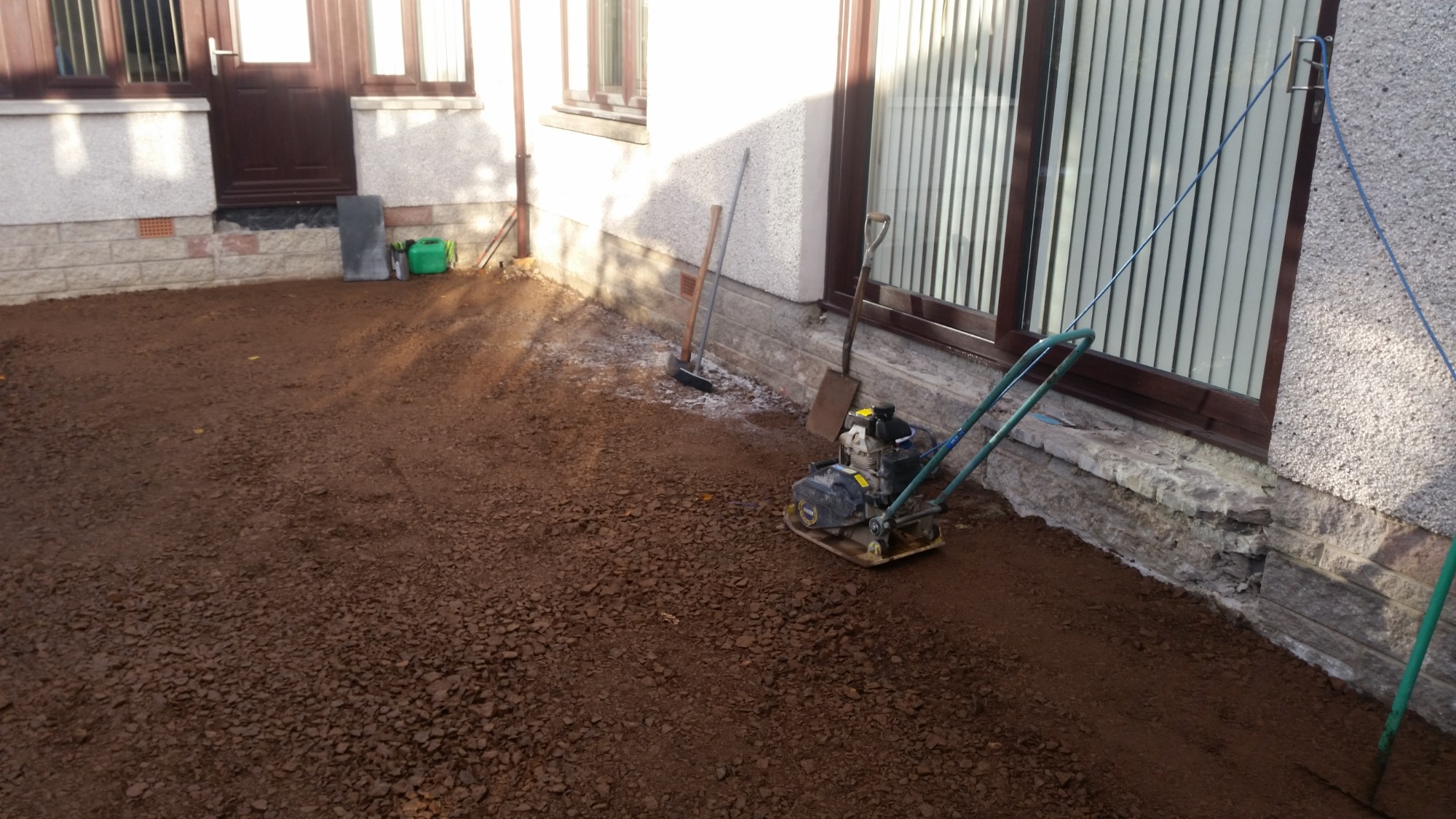Przygotowanie podłoża z gruzu i piasku pod kostkę brukową przy ścianie budynku, ubijarka wibracyjna, grabie i łopata w tle.