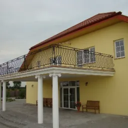 Ż&oacute;łty dom z balkonem z czarną, ozdobną balustradą i kolumnami w stylu greckim, widok z zewnątrz, z elementami architektury klasycznej.