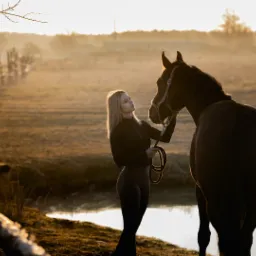 Kobieta w czarnym stroju głaszcze konia na tle mglistego, złotego krajobrazu z drewnianym płotem i małym stawem.