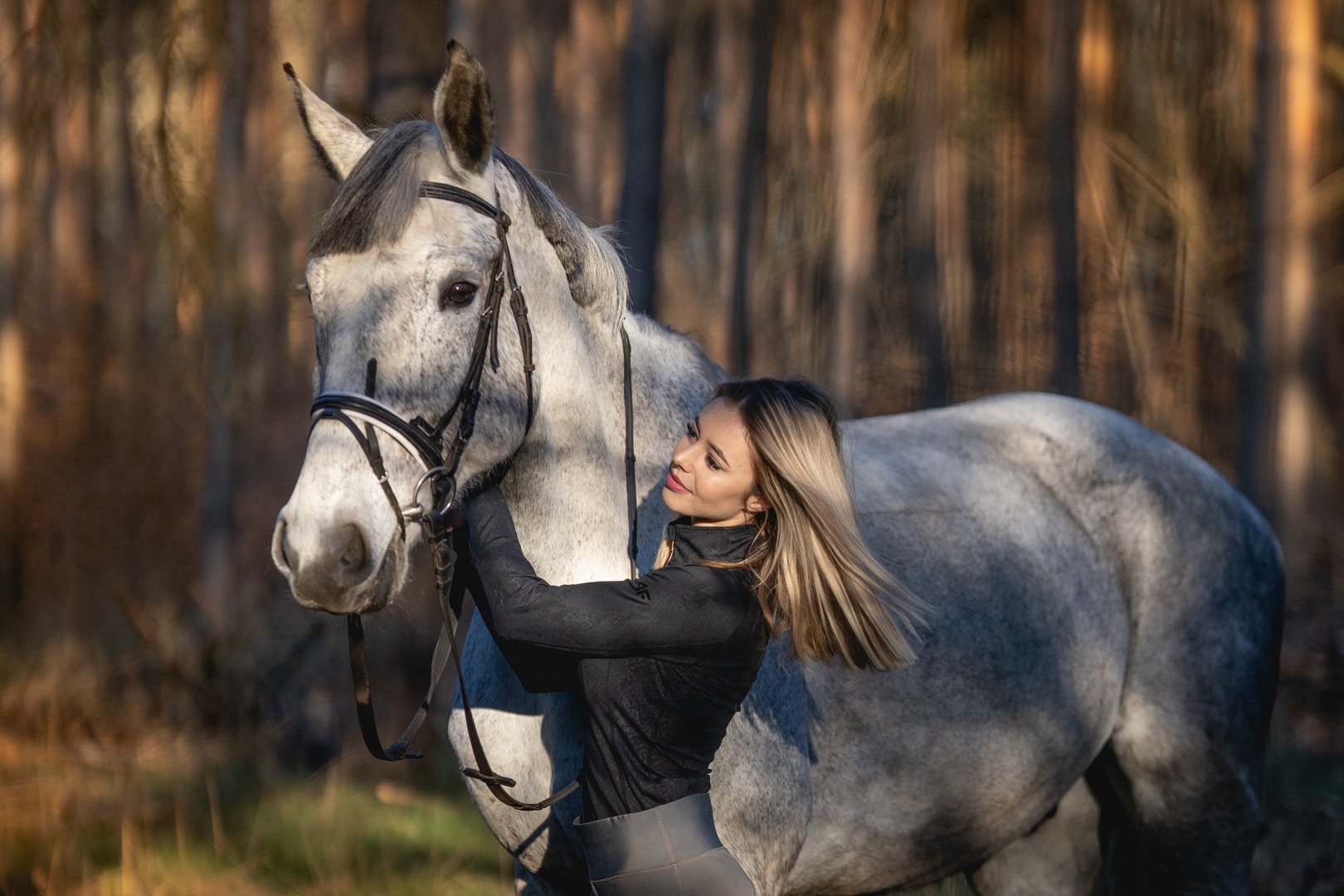 Młoda kobieta w czarnej bluzce i szarych spodniach obejmuje pysk siwego konia w uprzęży na tle lasu w ciepłym świetle popołudniowego słońca.