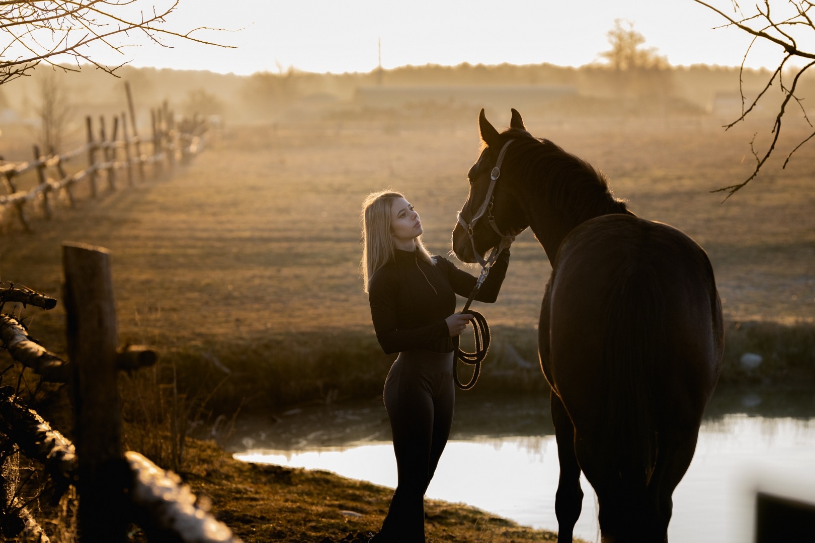 Kobieta w czarnym stroju głaszcze konia na tle mglistego, złotego krajobrazu z drewnianym płotem i małym stawem.