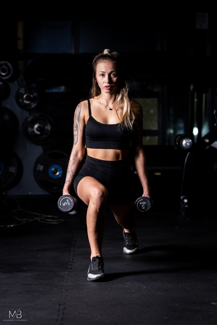 Kobieta w sportowym stroju wykonuje wypad z hantlami w siłowni, widoczne tatuaże na ramieniu i ciężary z napisem Aplus Sports 5 kg.