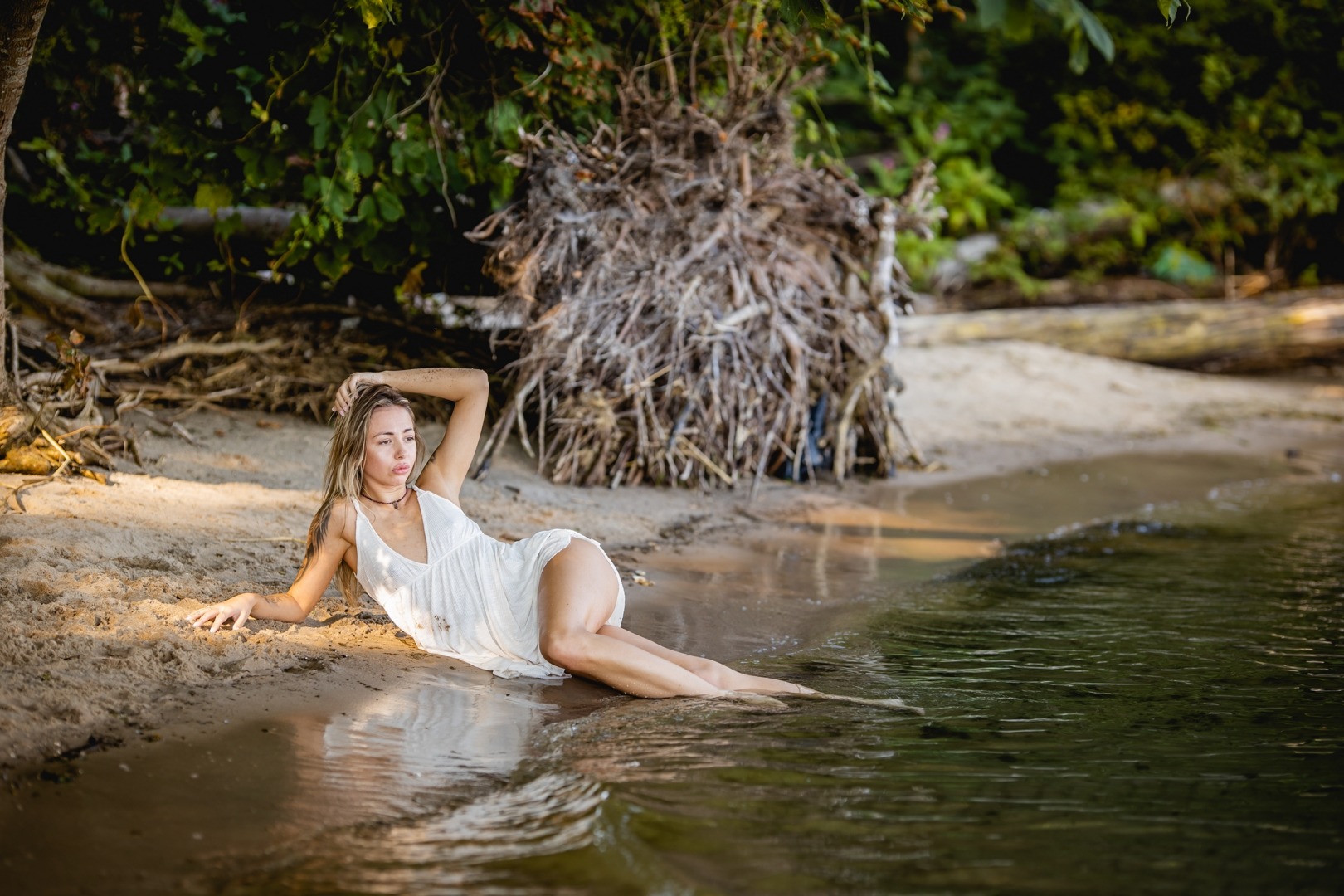 Kobieta w białej sukience leży na piaszczystej plaży, z nogami zanurzonymi w wodzie, zarośla w tle, delikatne oświetlenie.
