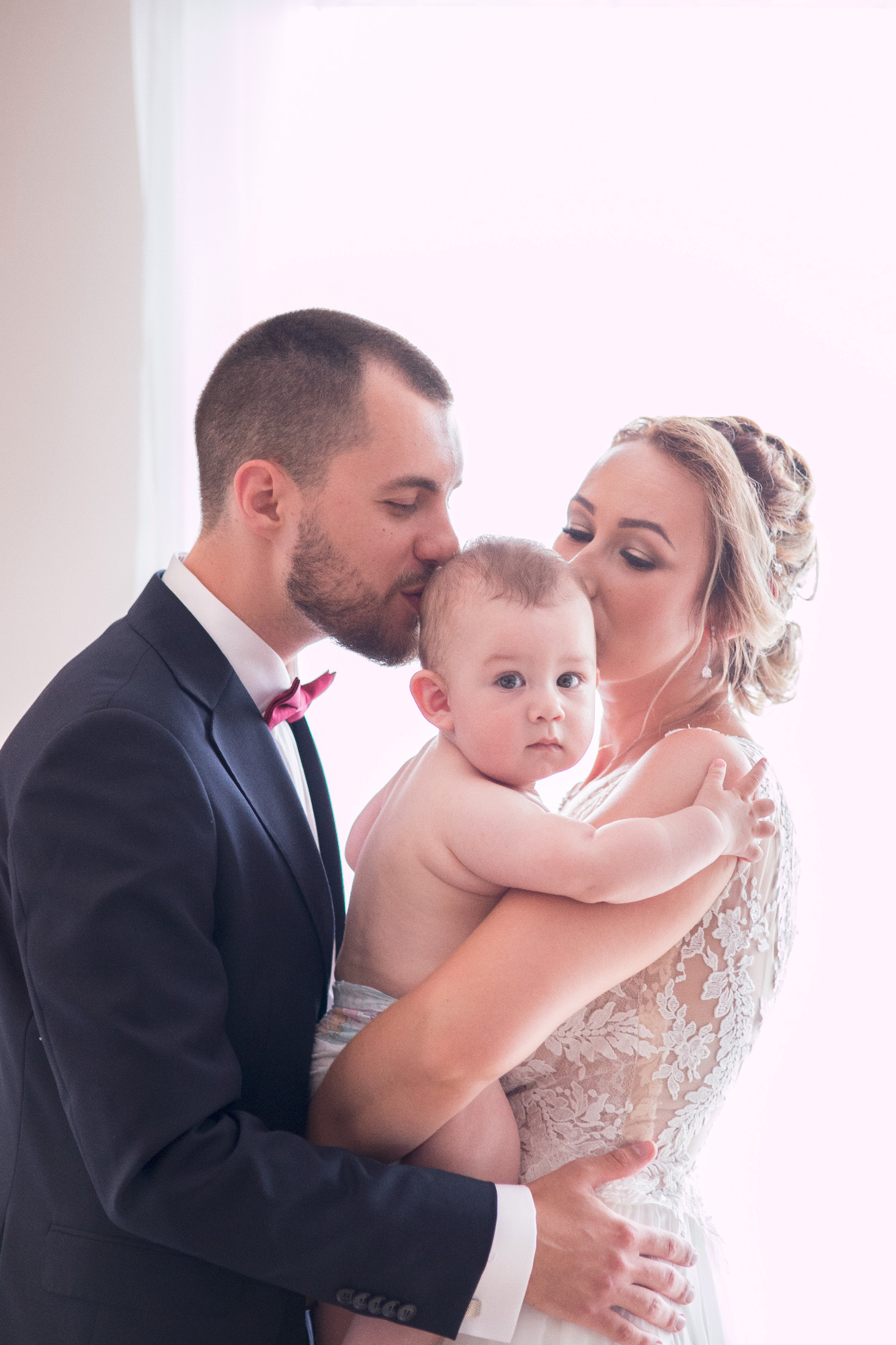 Elegancko ubrana para, panna młoda w koronkowej sukni ślubnej, trzyma niemowlę w pieluszce, oboje rodzice całują dziecko w głowę na jasnym tle.