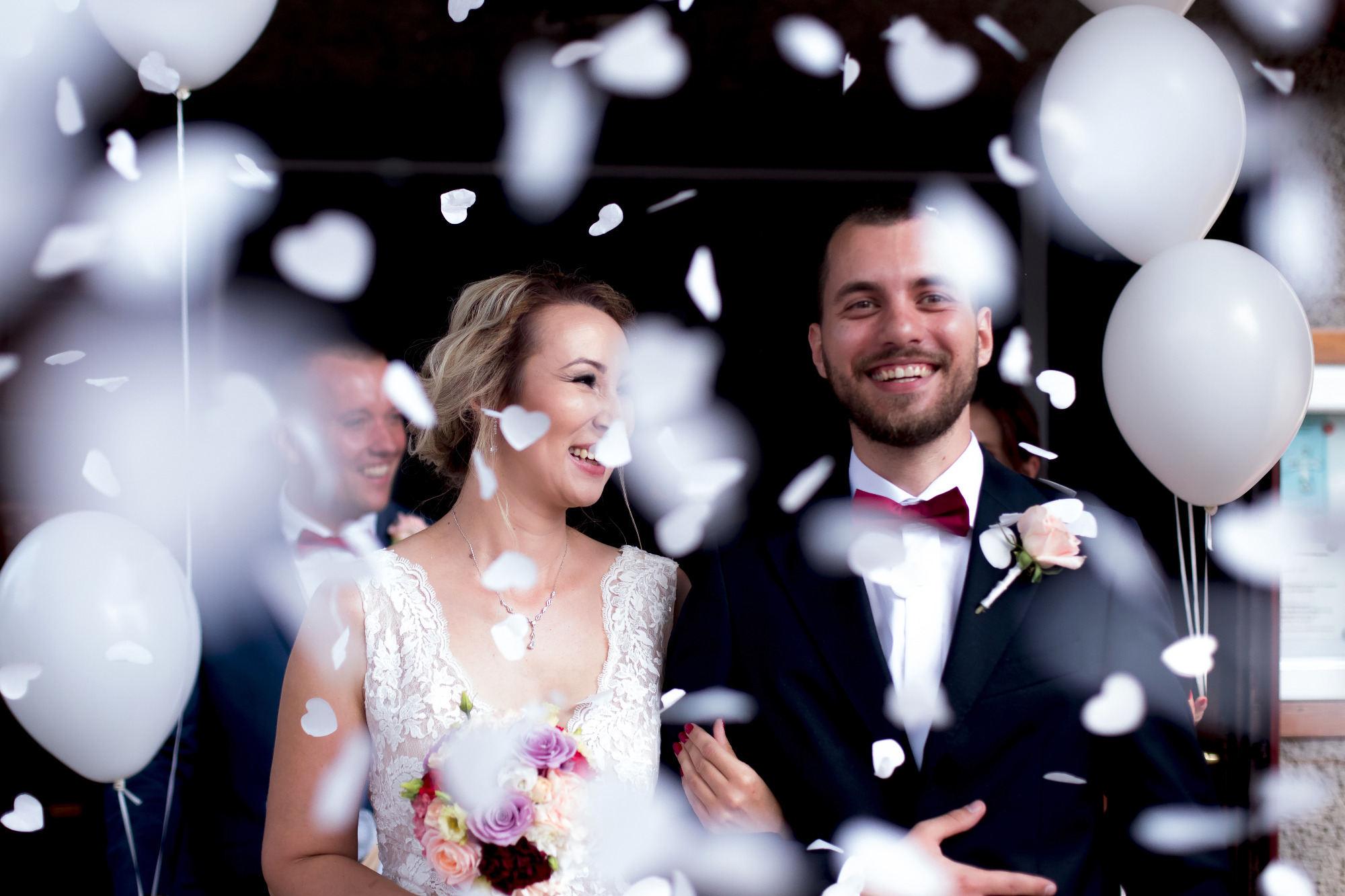 Uśmiechnięta para młoda wychodzi z kościoła obsypywana konfetti w kształcie serc, trzymając białe balony.