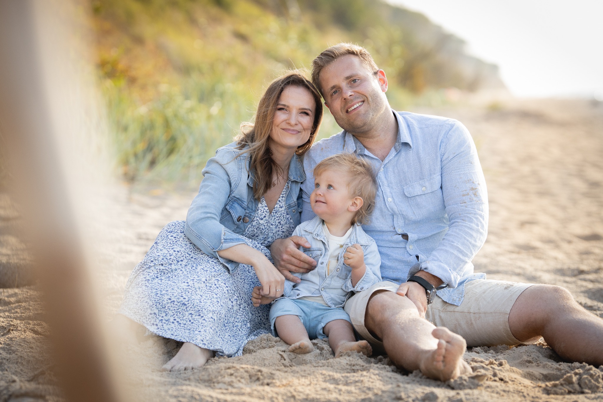 Uśmiechnięta rodzina siedzi na piaszczystej plaży: mama w kwiecistej sukience i jeansowej kurtce, tata w jasnoniebieskiej koszuli i beżowych szortach, a mały chłopiec w jeansowej koszuli...