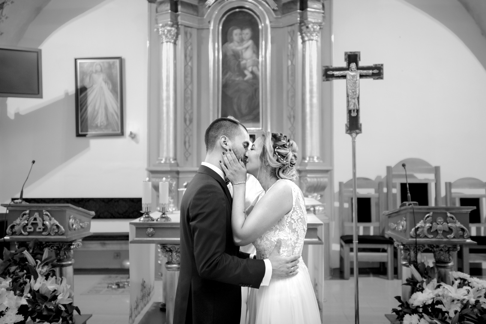 Czarno-białe zdjęcie całującej się pary młodej w kościele, z widocznym ołtarzem, obrazami religijnymi i kwiatami.