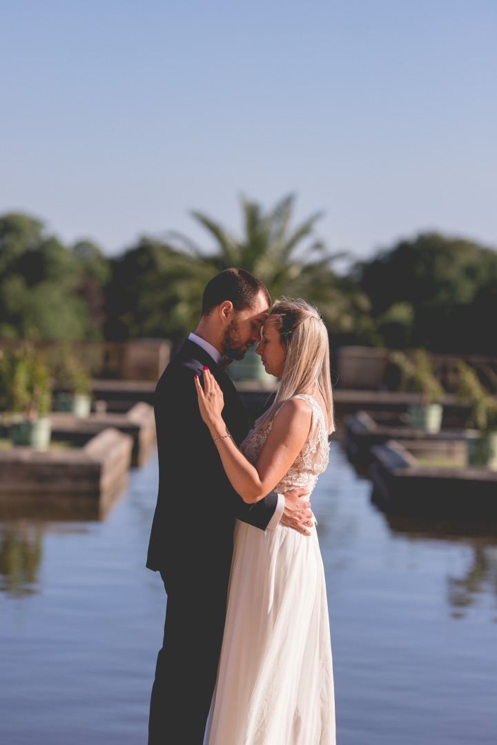 Para młoda w objęciach na tle wodnego ogrodu z palmami, ona w koronkowej sukni, on w granatowym garniturze.