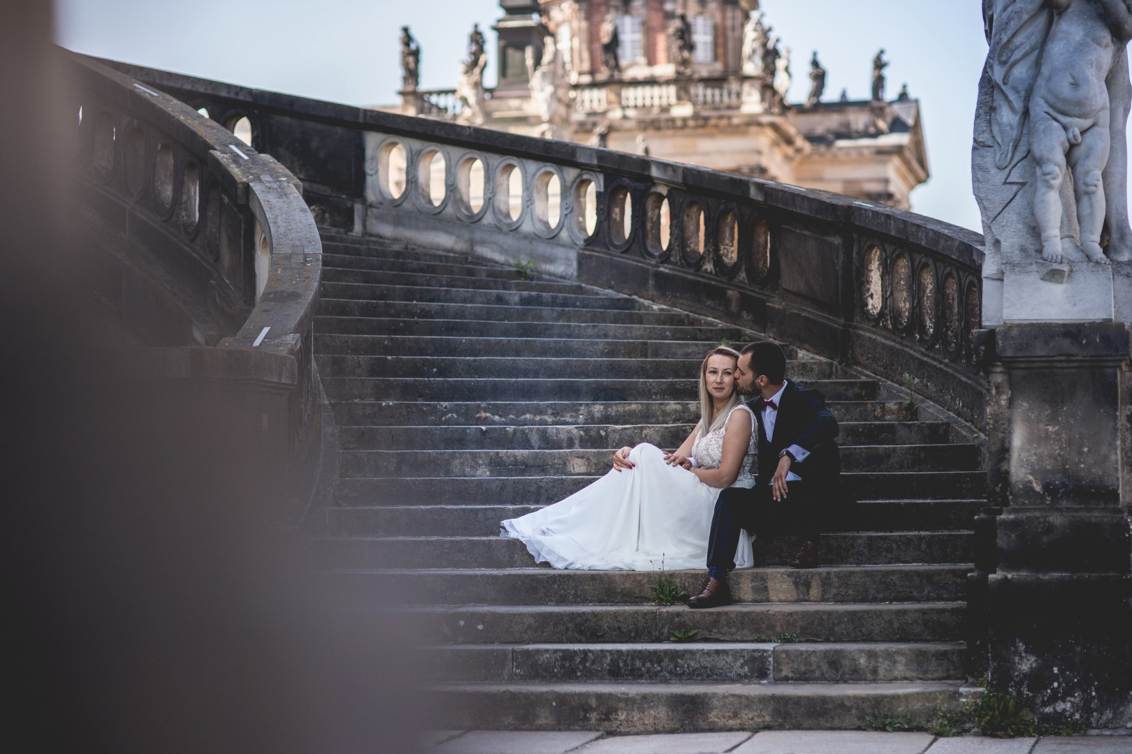Para młoda siedząca na kamiennych schodach zabytkowego budynku, mężczyzna całuje kobietę w policzek, w tle widoczna architektura z rzeźbami.