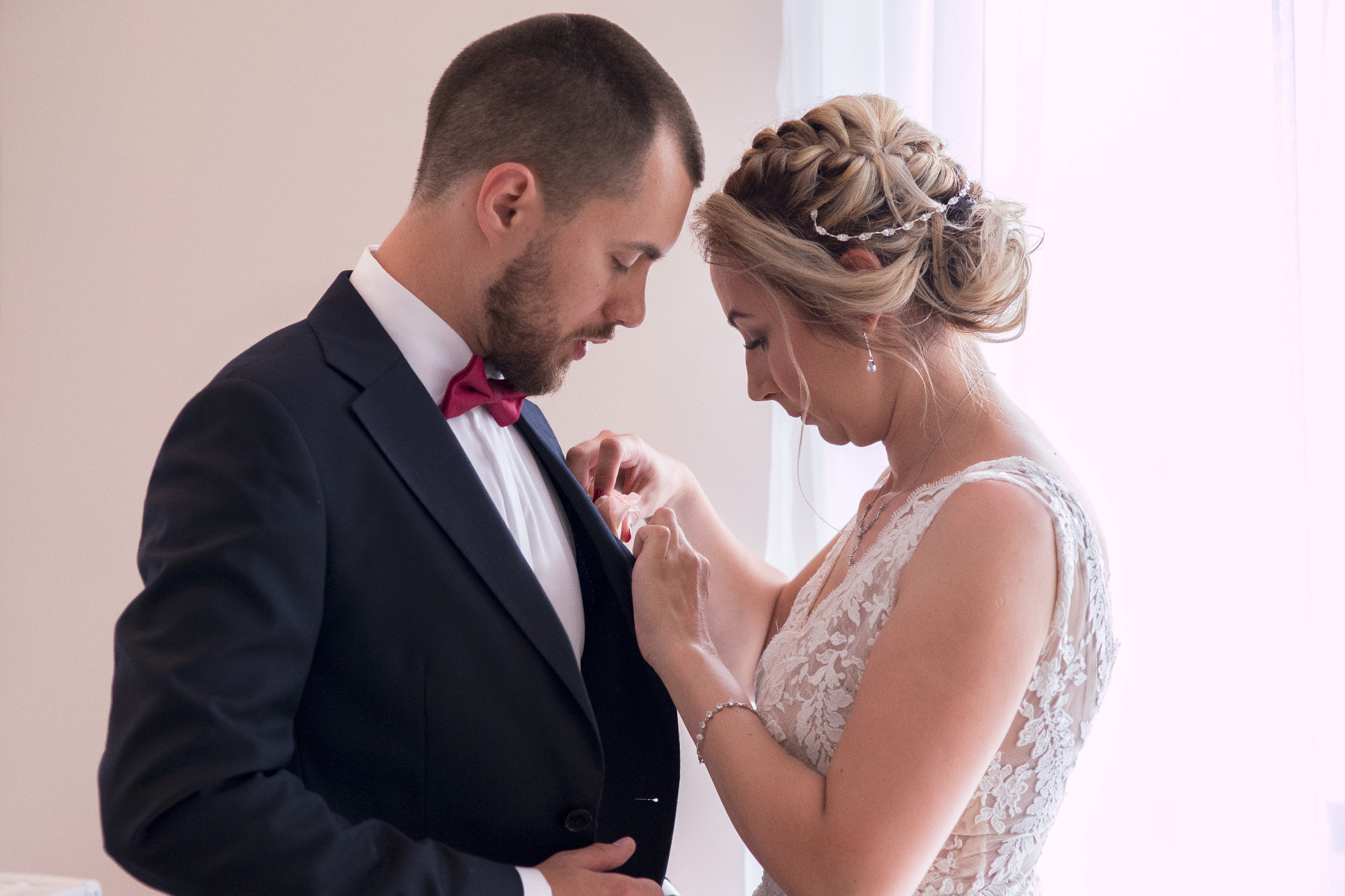 Panna młoda poprawia butonierkę pana młodego w eleganckim granatowym garniturze z bordową muchą, delikatna suknia ślubna z koronkowymi detalami, subtelny makijaż i upięcie włosów.