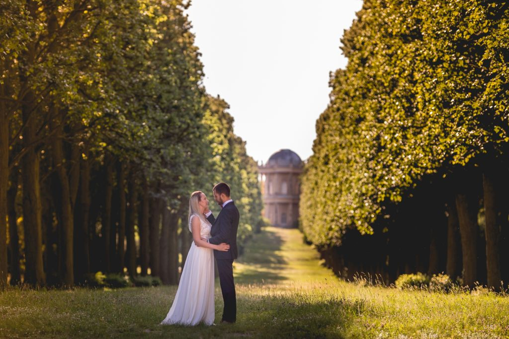Para młoda w objęciach na tle alei drzew prowadzącej do klasycystycznego budynku w słoneczny dzień.
