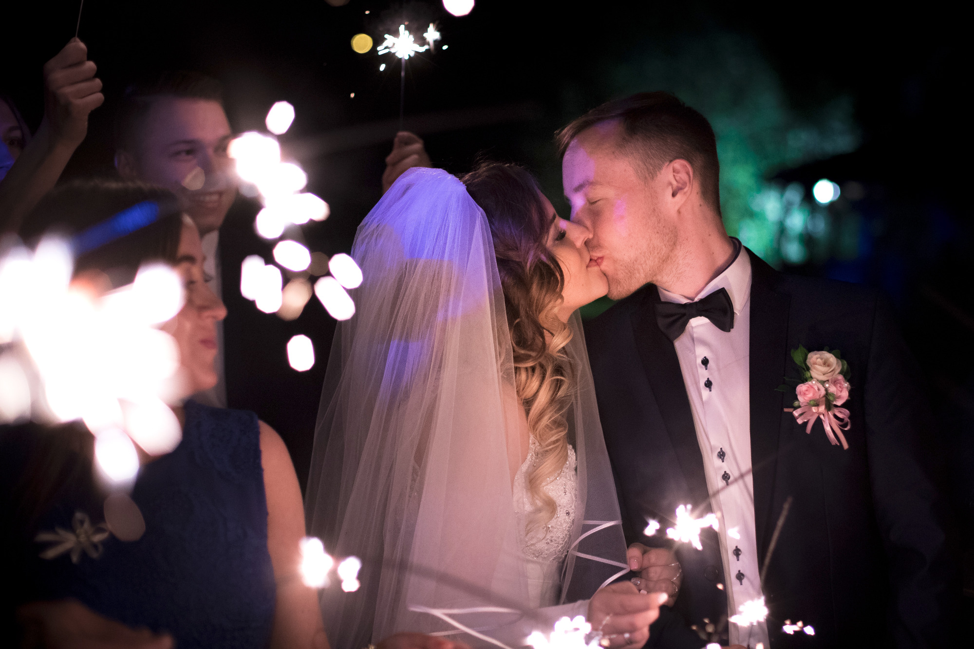 Pocałunek nowożeńców otoczonych przez gości z zimnymi ogniami w ciemną noc. Panna młoda w welonie, pan młody w smokingu z butonierką.