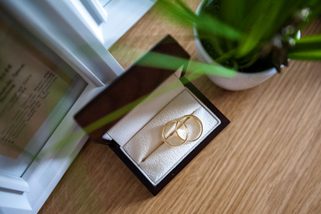 Złote obrączki w brązowym, otwartym pudełku na drewnianym stole, obok ramka ze starym tekstem i doniczka z zieloną rośliną.