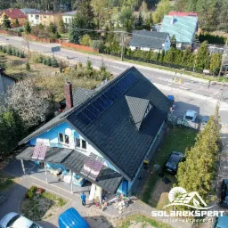 Widok z lotu ptaka na niebieski dom z zamontowanymi panelami słonecznymi na dachu i panelami kolektorów słonecznych nad wejściem, widoczny bałagan po instalacji i logo firmy w prawym dolnym rogu.