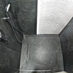 Wykończona kabina prysznicowa z ciemnoszarymi płytkami imitującymi kamień, chromowaną armaturą i odpływem liniowym, oraz białą ścianą z dekoracyjną strukturą fal.