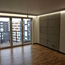 Wykończone wnętrze mieszkania z panelami dekoracyjnymi na ścianie, parkietem i dużym oknem balkonowym z widokiem na inne budynki.