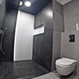 MAYKI Michał Ptaszek - Nowoczesna łazienka z kabiną prysznicową typu walk-in wykończona płytkami w odcieniach szarości i czerni, z białą ścianą z falistym wzorem i wiszącą toaletą.