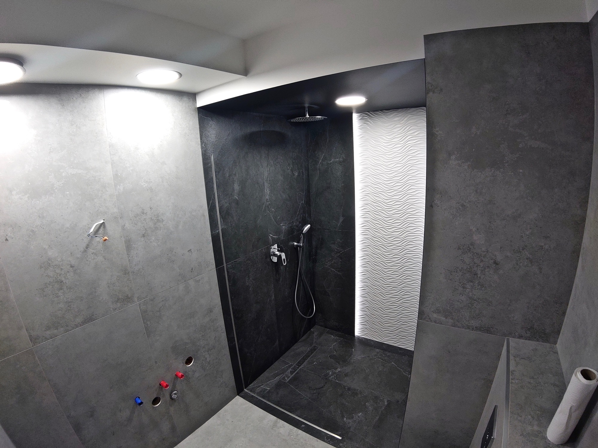 Nowoczesna kabina prysznicowa walk-in z szarymi płytkami imitującymi beton, czarnymi płytkami w strefie natrysku i dekoracyjną białą ścianą z falistym wzorem, oświetlona punktowymi lampami sufitowymi.