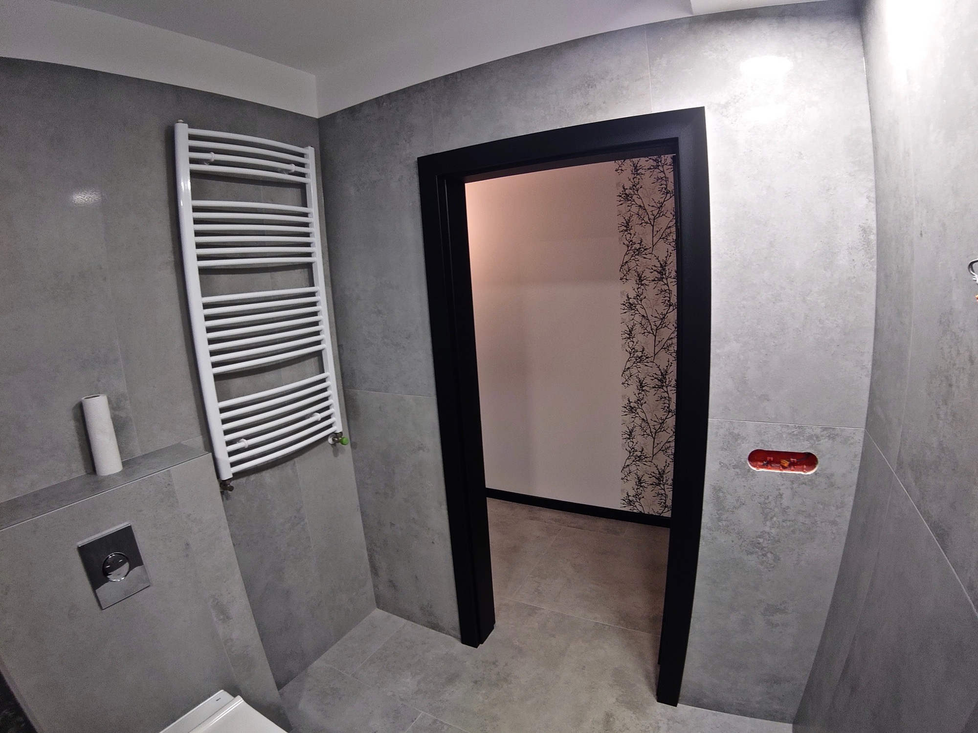 Szara łazienka z grzejnikiem drabinkowym, podwieszaną toaletą i czarnymi drzwiami prowadzącymi do pomieszczenia z tapetą w gałązki.