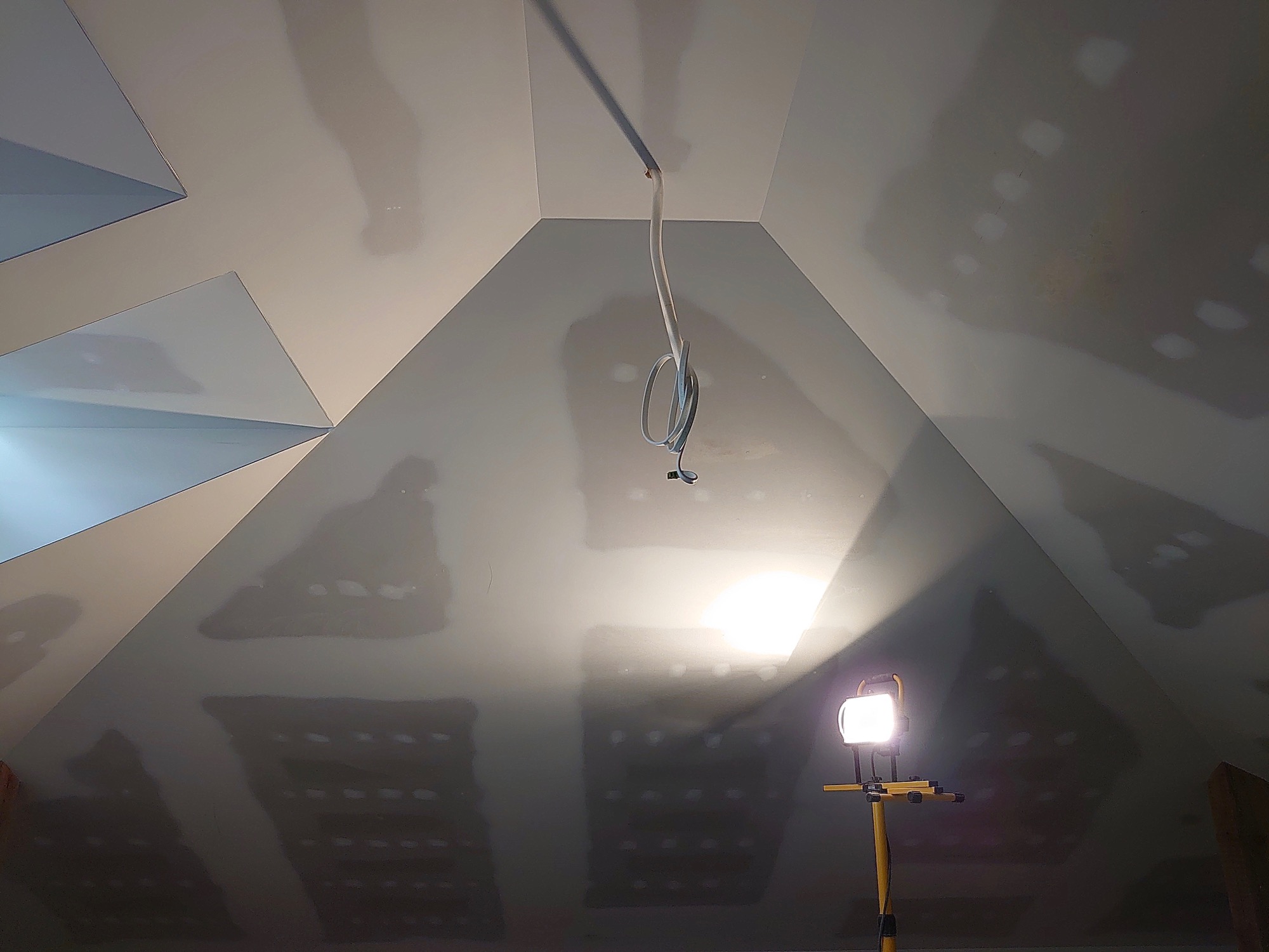 Sufit poddasza po szpachlowaniu we Wrocławiu, widoczne łączenia płyt gipsowo-kartonowych, kabel elektryczny zwisający z sufitu i lampa budowlana na statywie.