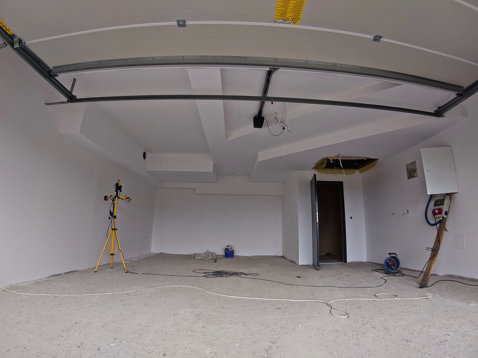 Wnętrze garażu po remoncie, świeżo pomalowane ściany na biało, widoczne przewody elektryczne i oświetlenie budowlane na statywie, brama garażowa pod sufitem.