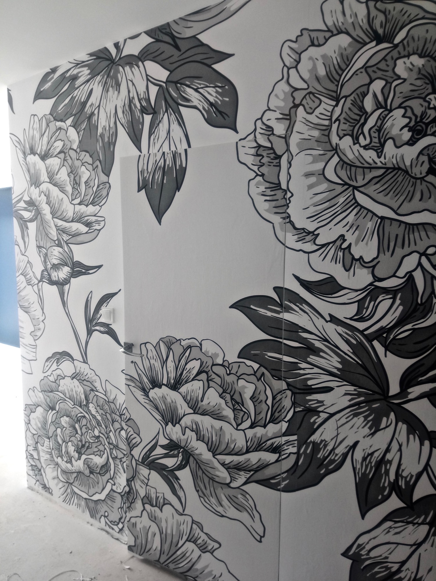 Ściana pokryta czarno-białym, ręcznie malowanym wzorem dużych kwiatów piwonii, obejmującym róg pokoju, z widocznym fragmentem niebieskiej ściany w tle.