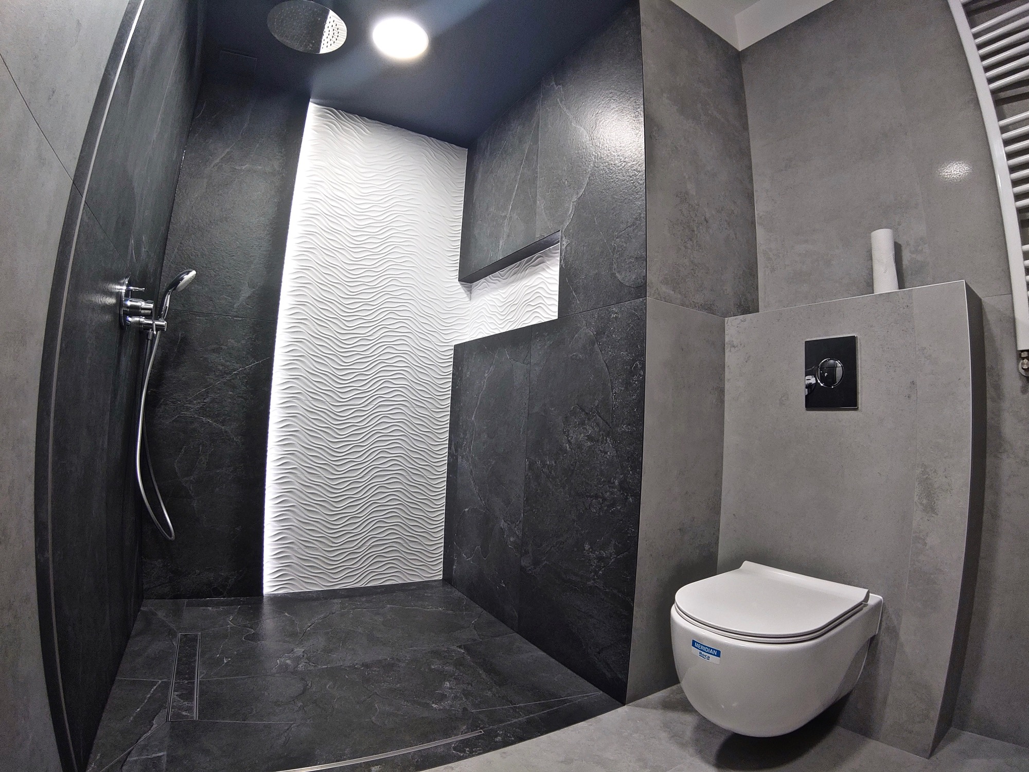 Nowoczesna łazienka z kabiną prysznicową typu walk-in wykończona płytkami w odcieniach szarości i czerni, z białą ścianą z falistym wzorem i wiszącą toaletą.