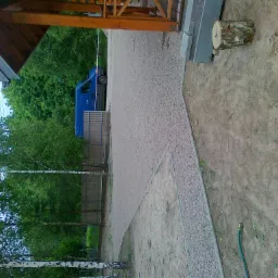 Żwirowa ścieżka ogrodowa z obrzeżem, fragment trawnika z wężem ogrodowym, pień drzewa, w tle zaparkowany niebieski bus i drzewa.