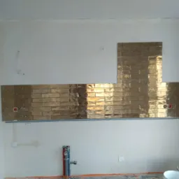 Fragment ściany z częściowo położonymi, błyszczącymi, złotymi płytkami w kształcie cegiełek. Widoczne otwory na gniazdka elektryczne i rury instalacyjne. Na dole ściany widoczne rury i wiadro.