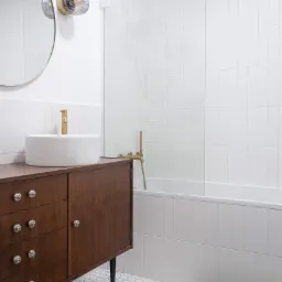 Nowoczesna łazienka z białymi, wąskimi płytkami ułożonymi pionowo, drewniana szafka pod umywalką z okrągłymi uchwytami, okrągłe lustro i złota armatura, podłoga wyłożona heksagonalnymi płytkami.