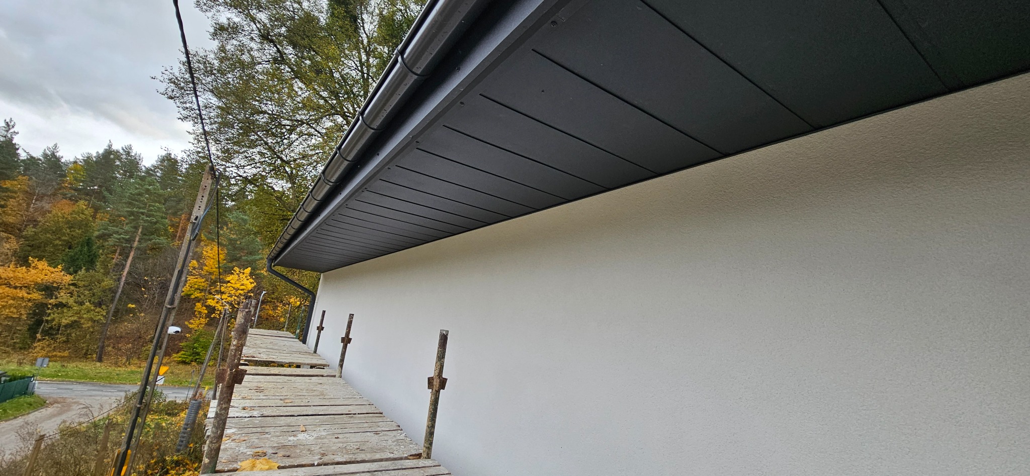 Szara podbitka dachowa z rynną, widok z rusztowania na tle jesiennego lasu. Ściana budynku o jasnej, ziarnistej fakturze. Ujęcie z dołu, podkreślające detale wykończenia.