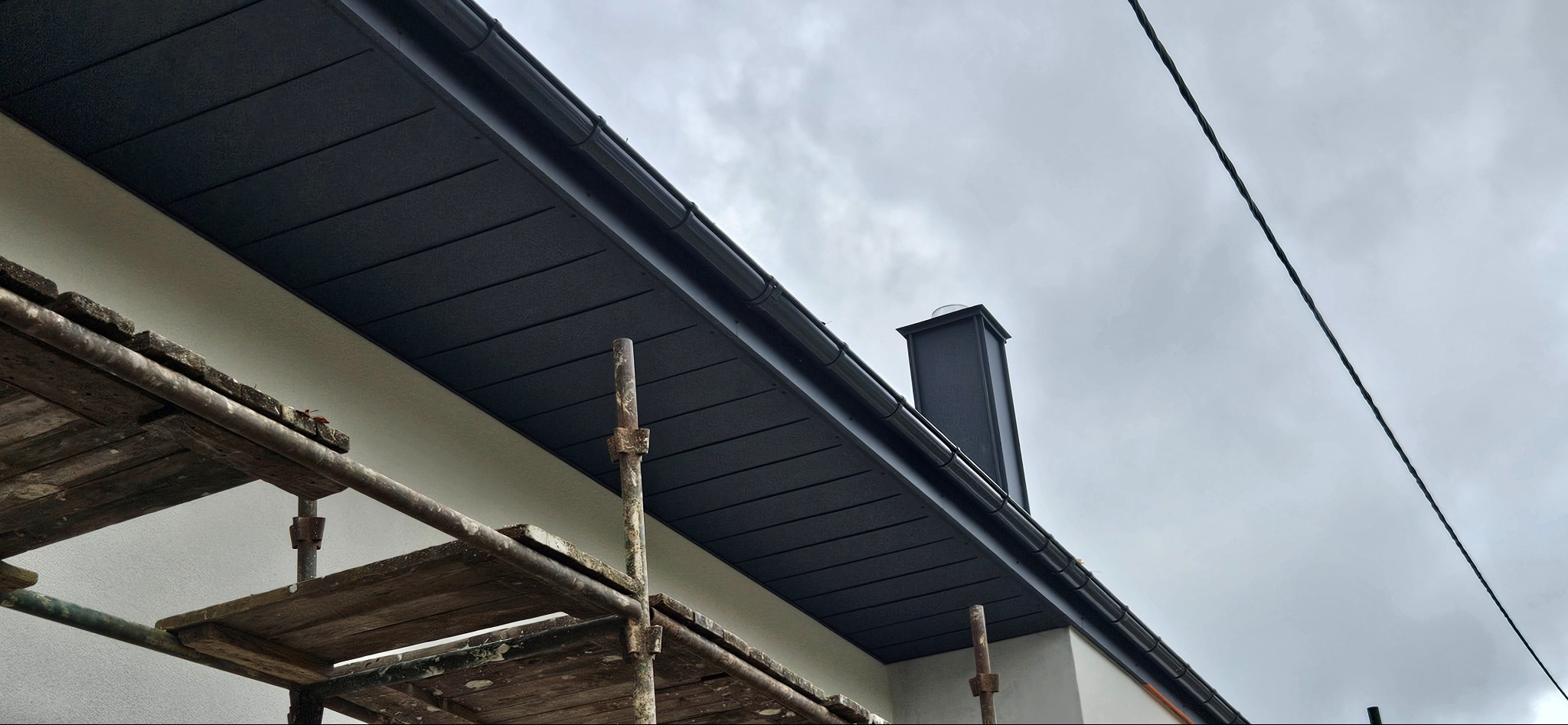 Ciemna podbitka dachowa z kominem, widok z rusztowania na tle pochmurnego nieba. Detal wykończenia dachu z bliska.