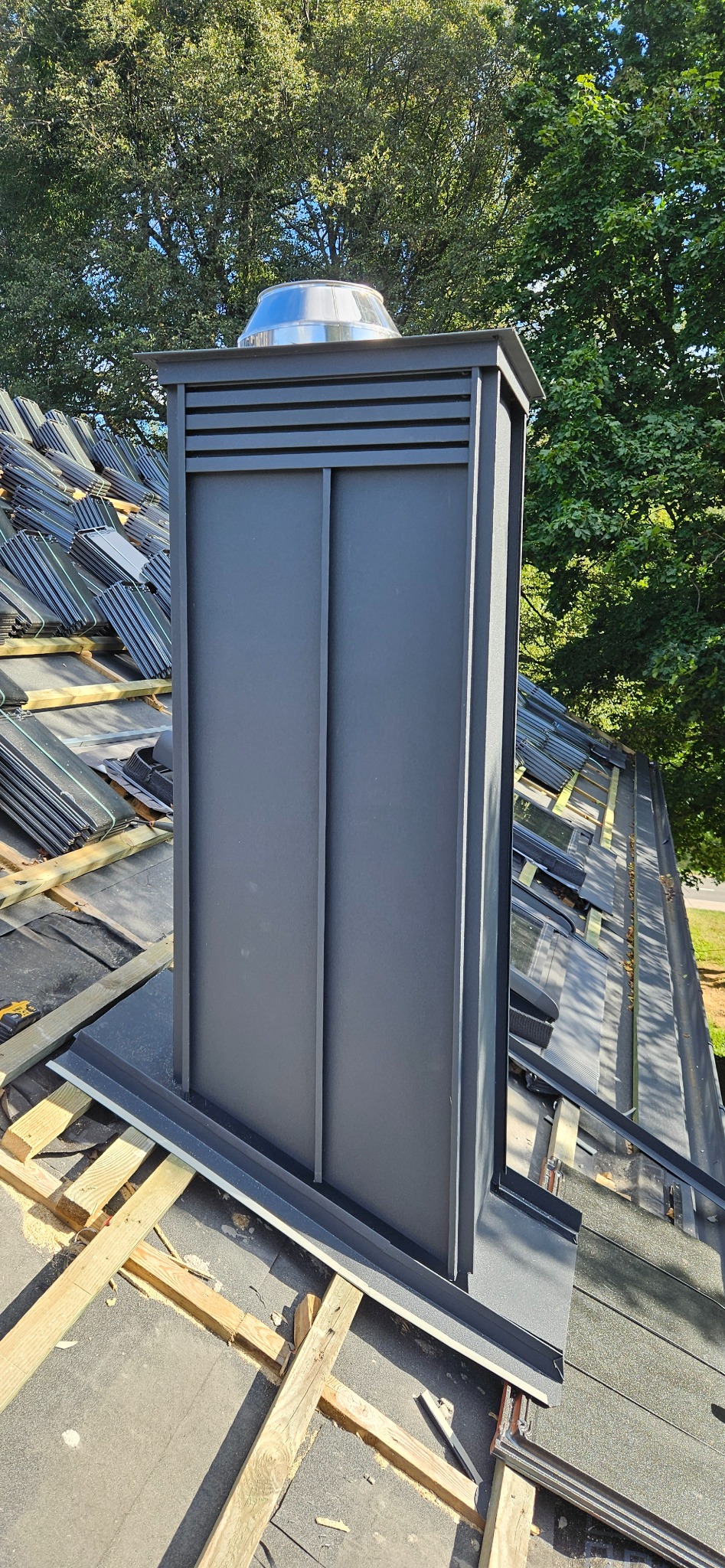 Szary komin wentylacyjny z metalowym daszkiem na dachu w trakcie krycia dachówką. Widoczne drewniane elementy konstrukcji dachu i zieleń drzew w tle.