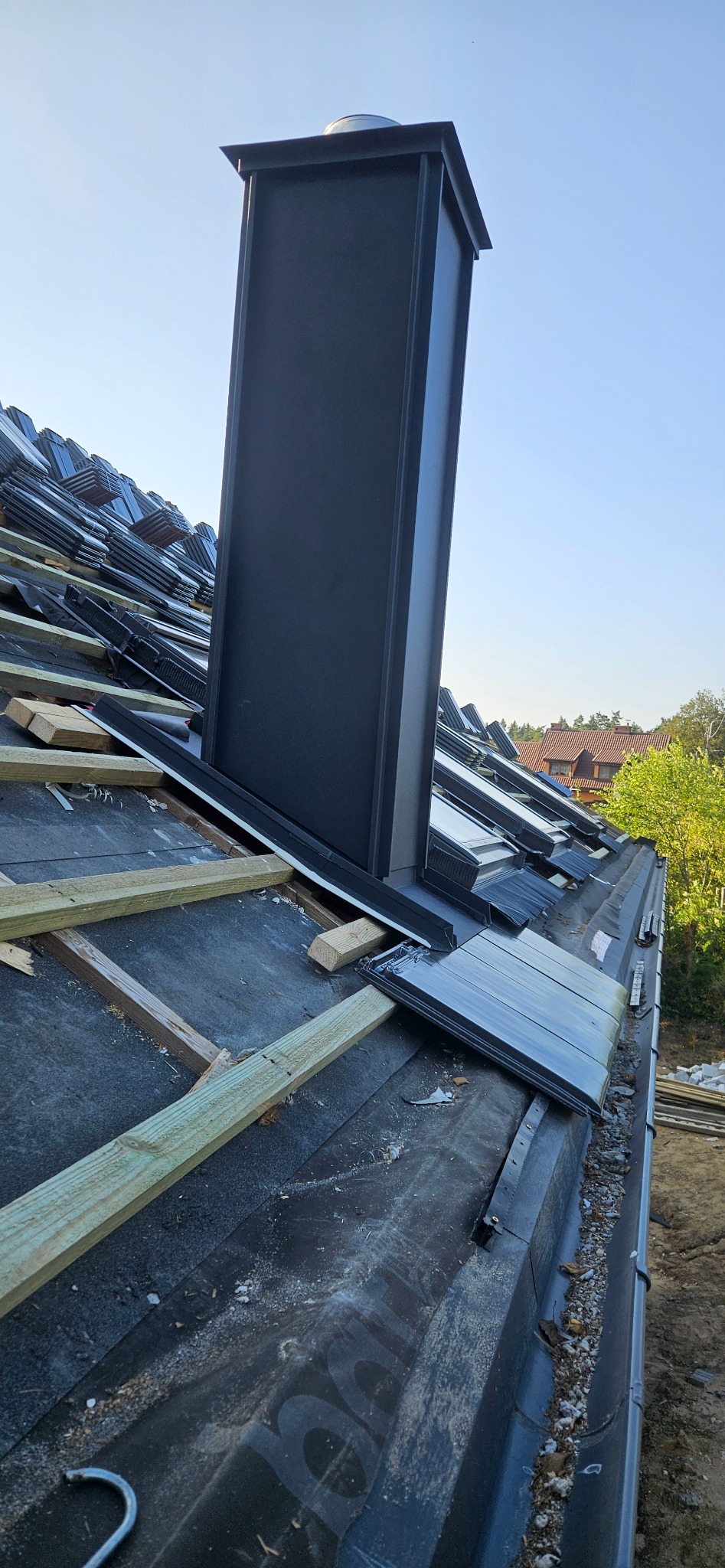 Dach z czarną membraną, drewnianymi łatami i kominem w trakcie krycia. Widoczne dachówki i okna dachowe. Ujęcie z dołu, w tle zieleń i zabudowania.