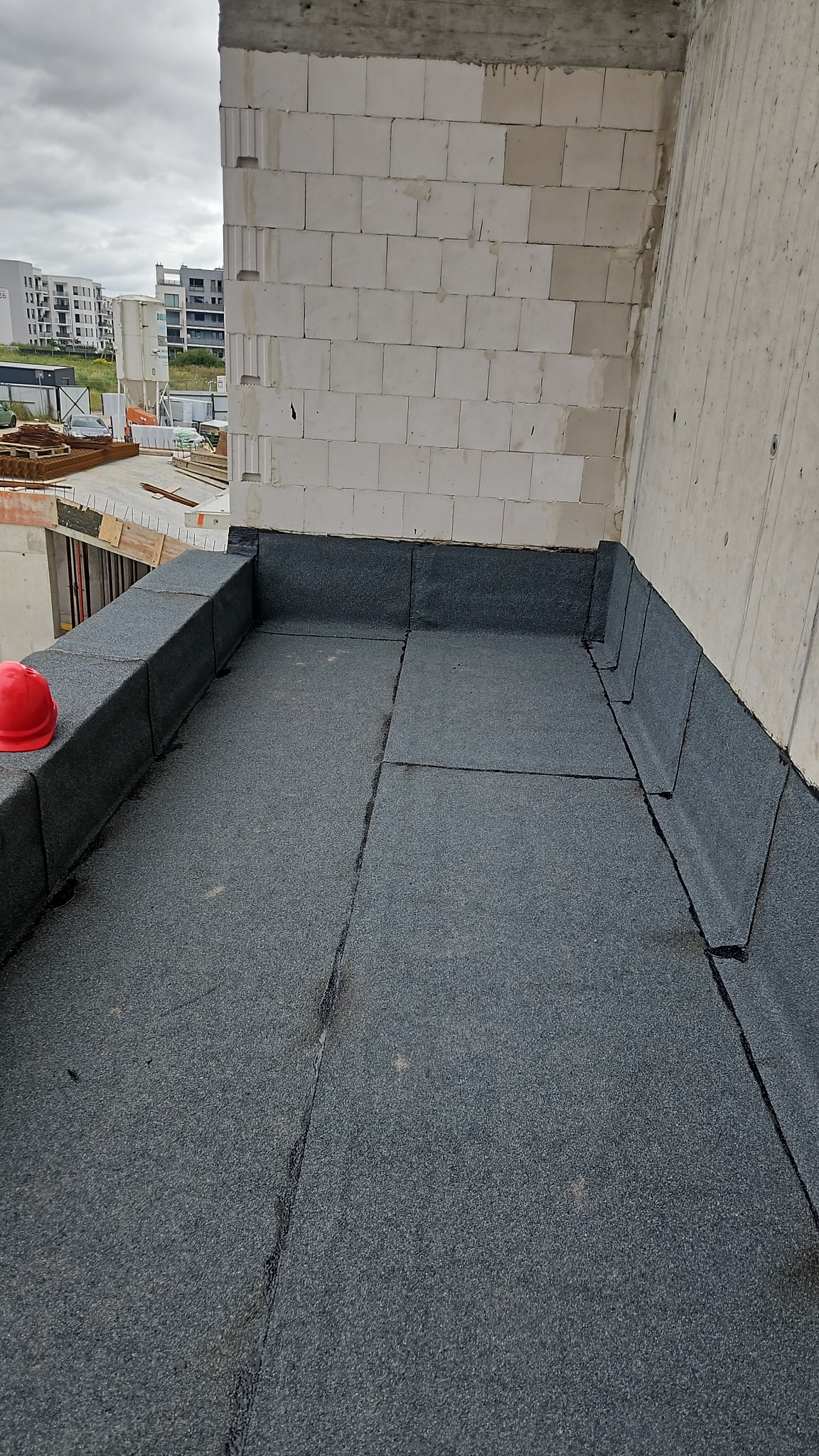 Szczelny, płaski dach pokryty papą termozgrzewalną na nowym budynku. Czerwony kask leży na krawędzi. Widok na miasto w tle.